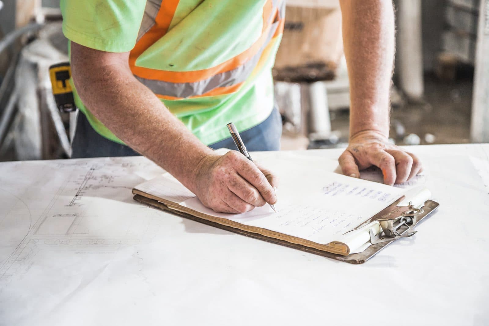 Site manager in a hi-vis vest writing notes on a clipboard over construction plans